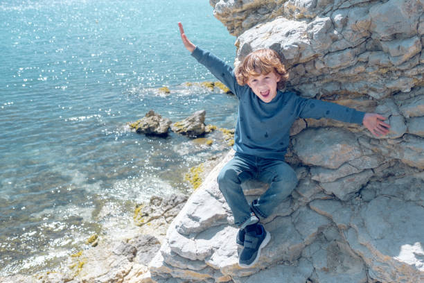 From above full body of positive boy with spread arms looking at camera while sitting on stony cliff above sea
