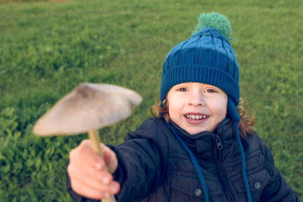 Delighted boy in warm outerwear and hat showing fresh picked mushroom in hand while standing on grassy field in nature