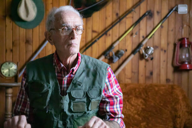 An elderly man in a plaid shirt and green utility vest sits at a wooden workbench, surrounded by tools and fishing equipment. The background features wooden paneling with fishing rods, a lantern, and a vintage clock, creating a rustic and outdoorsy atmosphere.