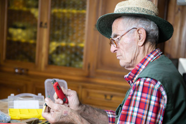 A senior man wearing glasses and a plaid shirt inspects a fishing reel at a workbench. Tools and accessories are scattered on the table, with a rustic workshop background. Perfect for themes of craftsmanship, hobbies, and senior activities.