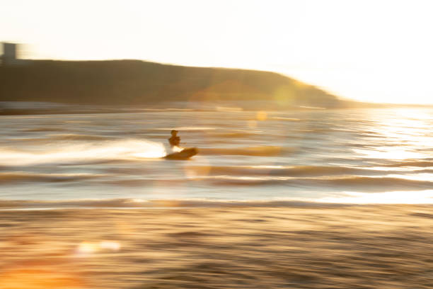 LENS FLARE, LOW ANGLE VIEW: Athletic surfer runs barefoot on the sand during sunset, carrying his surfboard. Young man is rushing towards the ocean to catch some waves before the golden sun sets.