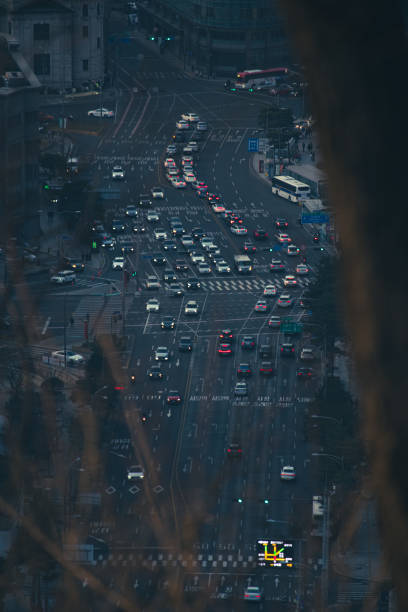 The image shows a view of a busy intersection in Seoul, captured from a high vantage point. The wide, multi-lane road is filled with vehicles, including cars, buses, and taxis, moving in various directions. The traffic is dense but orderly, typical of a bustling urban area. The scene is set during dusk or early evening, with the lights from the vehicles creating a line of red and white, highlighting the flow of traffic. Surrounding the road are tall buildings, adding to the sense of a dense, modern cityscape. The overall atmosphere reflects the dynamic and fast-paced nature of life in Seoul.