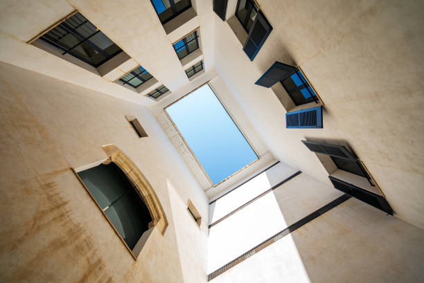 This upward view through an open courtyard in Palma de Mallorca, Spain, reveals a clear blue sky framed by the white walls of the surrounding buildings. The open windows and the geometric lines create a striking composition, highlighting the modern elements within a traditional Mediterranean setting.