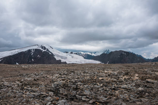 istock Dramatic misty top view from stone pass to big glacier tongue among sharp rocks and large snow-capped pinnacle in rainy low clouds. Dark atmospheric mountain silhouettes in rain in gray cloudy sky. 2169802083