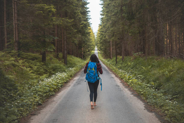 istock Back view of a woman with a blue backpack walking barefoot down a peaceful forest road, surrounded by lush greenery and tall trees, symbolizing adventure and tranquility in nature 2169809361