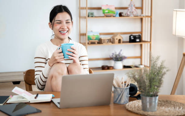 istock Young smiling happy beautiful asian woman relaxing using laptop computer and drinking coffee in the living room at home. 2169810511