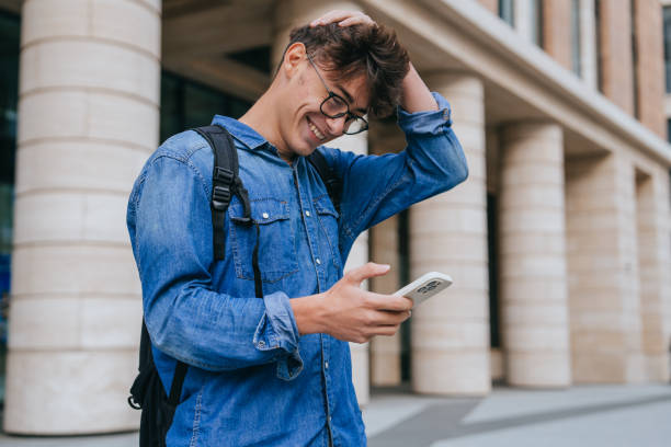 istock young man in a denim shirt, smiling while using his smartphone and carrying a backpack in a modern city environment. Concept of digital communication and urban lifestyle 2169816482