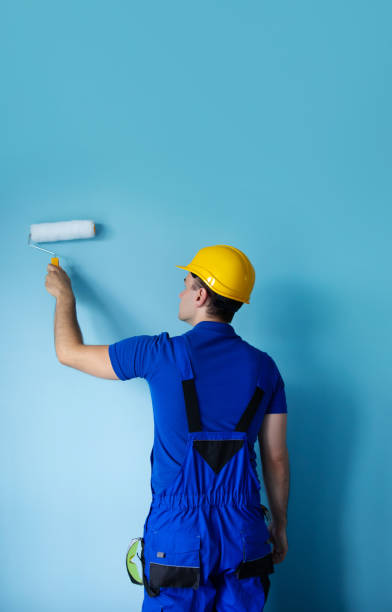 istock A construction worker in blue overalls and a yellow hard hat paints a wall with a roller. The image captures the worker's back as he methodically applies paint, demonstrating attention to detail. 2169836845