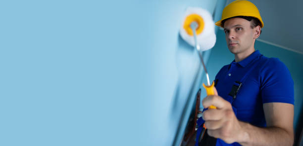 A construction worker in a yellow hard hat and blue overalls carefully paints a wall with a roller. The close-up shot captures his focus and precision, emphasizing the meticulous nature of his work.