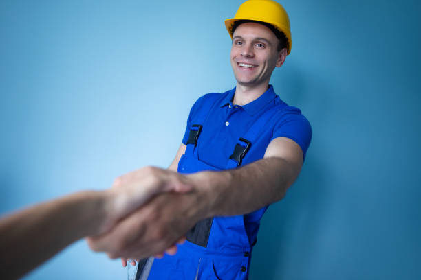 istock A smiling builder in blue overalls and a yellow hard hat shakes hands, representing successful collaboration. The background is a clean blue wall, highlighting the workers professionalism 2169837105