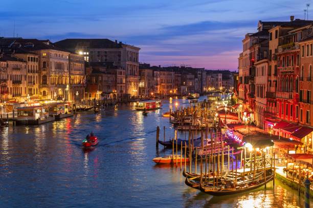 Night ambiance at Venice’s Grand Canal featuring shimmering reflections, illuminated classic architecture, and moored boats along its famous waterway. The serene scene exudes timeless charm and cultural significance.