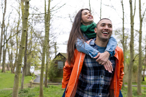 Medium shot of a husband and wife together in a woodland area next to a luxury glamping pod they are staying at located in Northumberland, Northeastern England. The woman is piggybacking on his back. They are both smiling and looking away from the camera.\n\nVideos are available similar to this scenario.