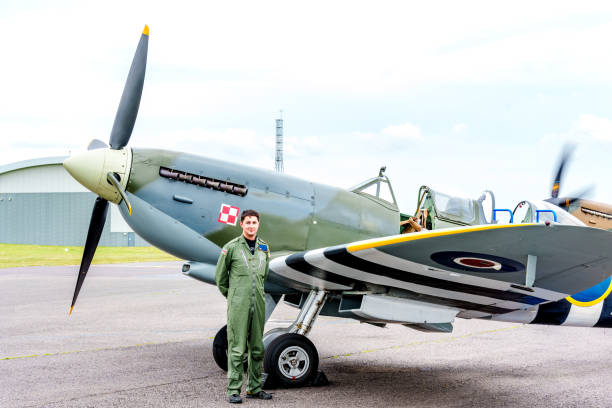 a man dressed in a green flight suit standing next to a historic aircraft,  a World War II-era Spitfire, on the tarmac outside a building. The aircraft features its characteristic propeller and markings, including a Polish Air Force insignia. The setting appears to be an aviation museum or airfield, with another similar aircraft visible in the background. The scene evokes a sense of history and aviation heritage, with the man possibly being a pilot , adding a personal element to this glimpse of iconic aviation history.