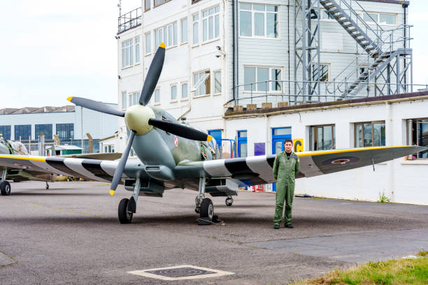 a man dressed in a green flight suit standing next to a historic aircraft,  a World War II-era Spitfire, on the tarmac outside a building. The aircraft features its characteristic propeller and markings, including a Polish Air Force insignia. The setting appears to be an aviation museum or airfield, with another similar aircraft visible in the background. The scene evokes a sense of history and aviation heritage, with the man possibly being a pilot , adding a personal element to this glimpse of iconic aviation history.