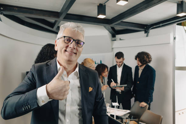 istock Senior businessman gesturing thumbs up with team working in background 2169877565