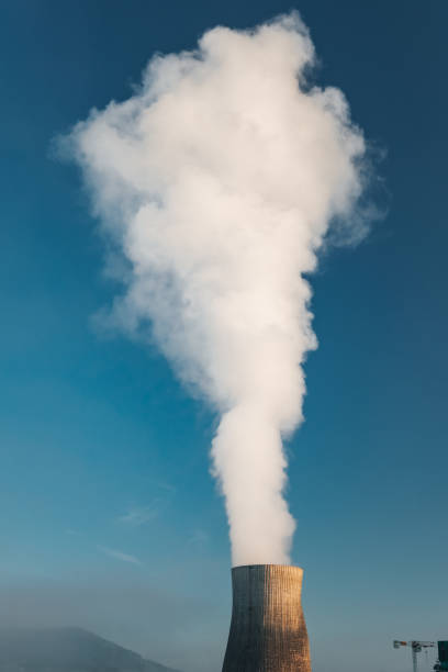Image of a power plant at sunrise, featuring two large industrial chimneys emitting smoke into the clear blue sky. This photo is ideal for use in topics related to energy production, environmental impact, and industrial landscapes