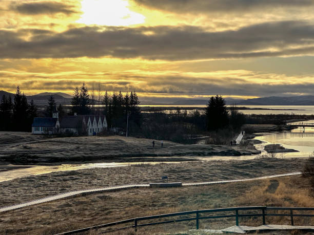 A serene landscape with a small, charming house surrounded by trees, a boardwalk path, and a tranquil body of water, all under a cloudy sky with the sun glowing through.