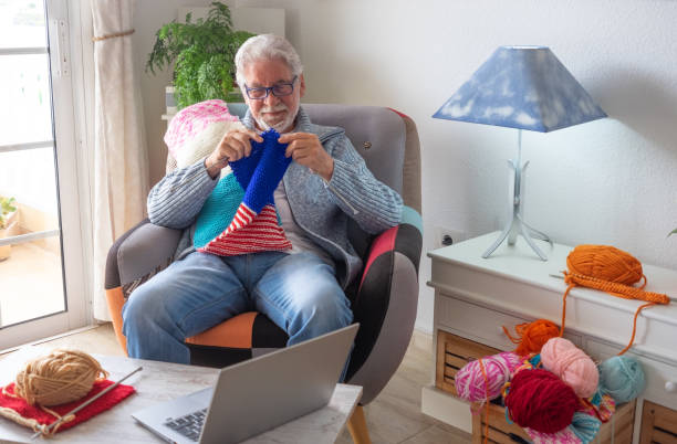 istock Senior man at home follows online knit tutorial to relax enjoying resting home leisure activity sitting on armchair - male people knitting with computer class help - people inner lifestyle hobby 2169936467
