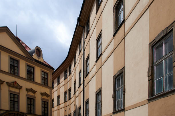 A view of historical buildings in Prague with beige and tan facades, featuring a curved section of the structure. The windows are framed by darker, aged stone, reflecting the city's architectural charm. The scene captures the quiet elegance of an alleyway between two buildings, with a cloudy sky above. The reddish-tiled roof on one of the buildings adds a classic Central European architectural element to the scene.