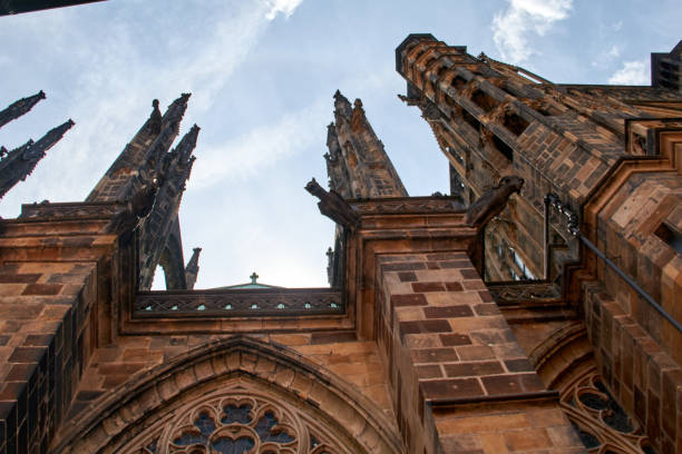 A close-up view of the towering Gothic spires of St. Vitus Cathedral, showcasing its intricate stonework and gargoyles. The shot is taken from a low angle, emphasizing the height and grandeur of the cathedral. The blue sky with light clouds forms a beautiful contrast with the dark tones of the stone structure, highlighting the architectural magnificence of this iconic landmark in Prague Castle.