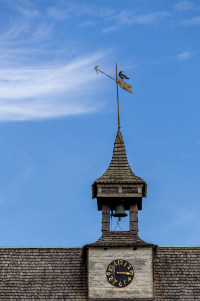 A close-up view of a clock tower with a weather vane on top, featuring a bell and a clock face. The sky is blue with wispy clouds in the background.