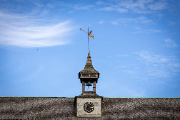 A close-up view of a clock tower with a weather vane on top, set against a blue sky with wispy clouds. The tower features a bell and a decorative spire.