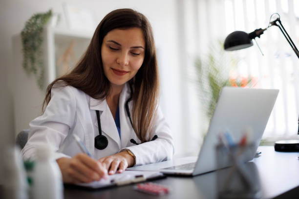 istock Smiling female doctor taking notes while working on laptop in the office 2169942774