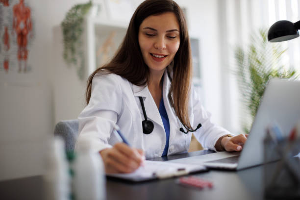 istock Smiling female doctor taking notes while working on laptop in the office 2169942780