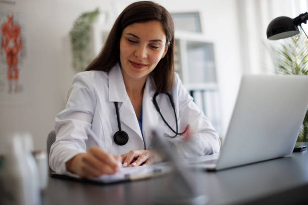 istock Smiling female doctor taking notes while working on laptop in the office 2169942781
