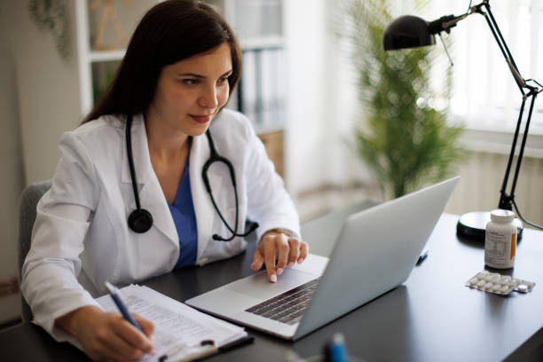 istock Smiling female doctor taking notes while working on laptop in the office 2169942812