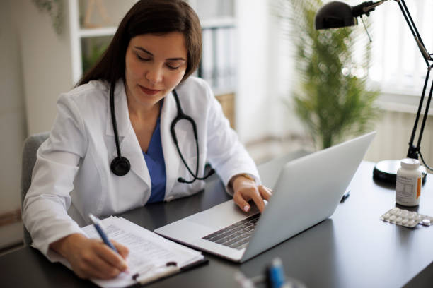 istock Smiling female doctor taking notes while working on laptop in the office 2169942841