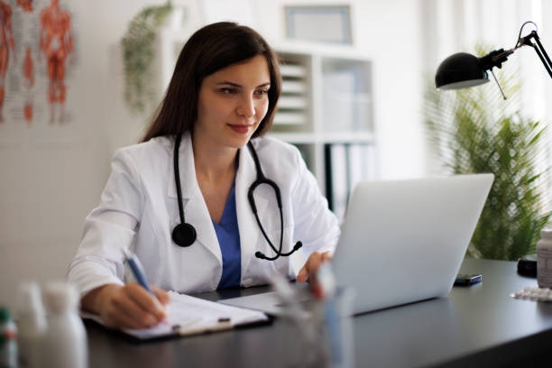 istock Smiling female doctor taking notes while working on laptop in the office 2169942842