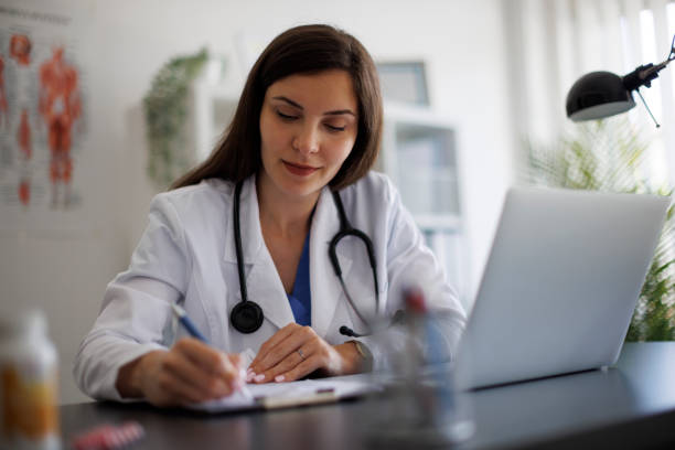 istock Smiling female doctor taking notes while working on laptop in the office 2169942875