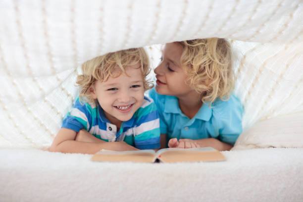 Child reading book in bed under knitted blanket. Two brothers playing together. Kids cozy bedroom hygge style. Little boy doing homework before sleep.