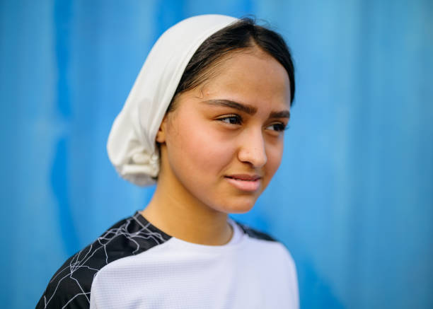 Female Soccer player wearing white hijab. Girl is against blue wall. She is wearing sports jersey.