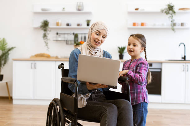 istock Mother in wheelchair with daughter using laptop in modern kitchen 2169973411