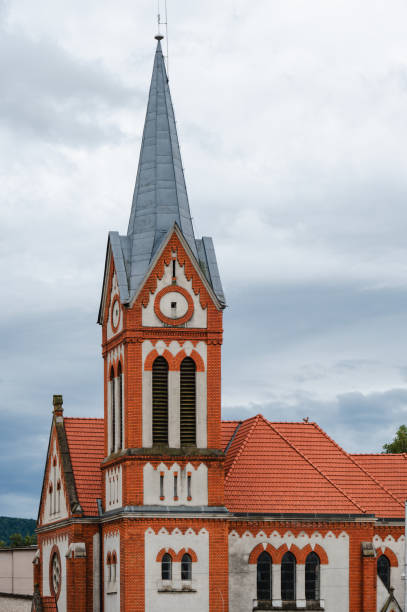 A detailed view of a Gothic Revival church featuring a tall, pointed steeple and a red brick facade. The church's architectural design includes arched windows and intricate detailing, set against an overcast sky, highlighting its historic and cultural significance.