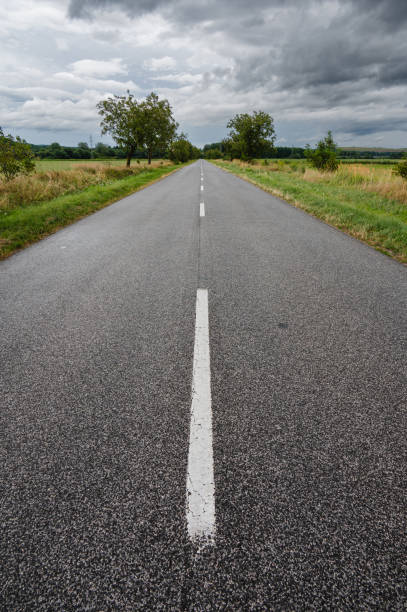 A straight, empty rural road stretches into the distance, lined with green grass and trees on both sides. The road leads towards a small village, with distant hills and mountains providing a scenic backdrop under a clear sky.