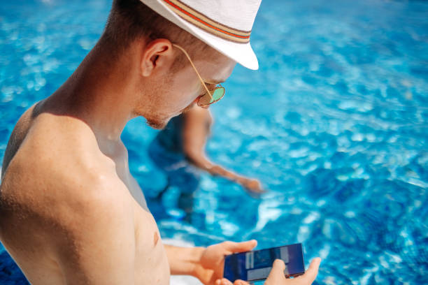 Young adult wearing hat and sunglasses standing in swimming pool while using smart phone