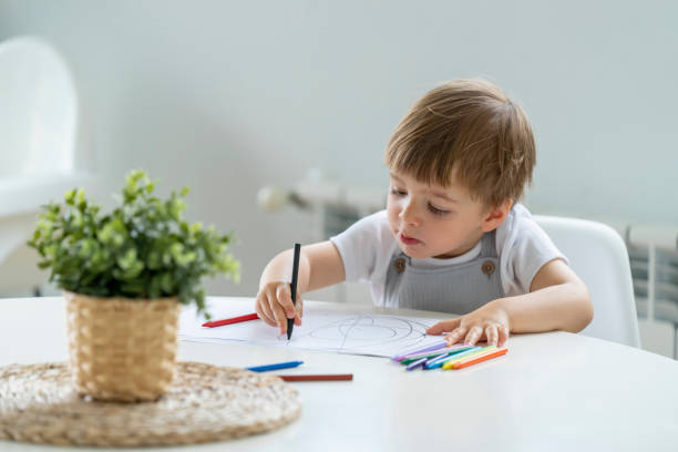 istock Little Boy Holding Two Crayons with a Pensive Look, Surrounded by Drawing Materials 2170002794