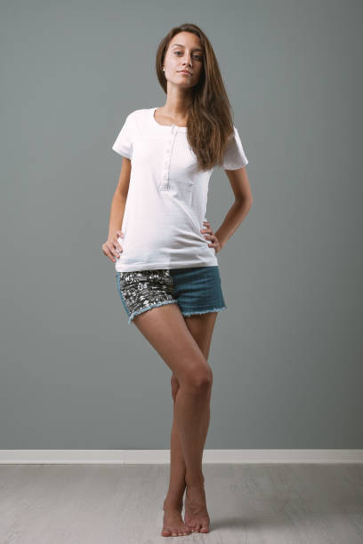 Young woman is standing with her hands on her hips and a neutral expression on her face. She is wearing jean shorts and a white t shirt