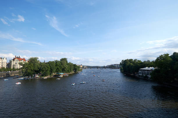 Prague, Czech Republic: A wide view of the Vltava River filled with small boats as people enjoy a peaceful day on the water. To the left, a historic building and trees line the riverbank, while on the right, more greenery and a riverfront café can be seen. The clear blue sky and calm river create a tranquil atmosphere, with a bridge visible in the distance connecting both sides of the city.