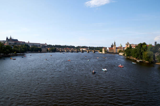 Prague, Czech Republic : A wide view of the Vltava River with boats scattered across the water, offering recreational rides to tourists. In the background, the iconic Charles Bridge connects both sides of the city, while the Prague Castle stands majestically on the left. The Old Town towers are visible on the right side of the riverbank, enhancing the city's historical charm. The bright summer sky and the calm waters reflect the peaceful yet vibrant atmosphere of Prague.