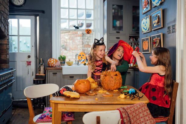 A medium side view of a mother carving pumpkins with her two daughters in their kitchen dressed in Halloween costumes. They are getting ready for Halloween and to go trick or treating locally. The mother is showing her how to carve the inside of the pumpkin out which is messy.