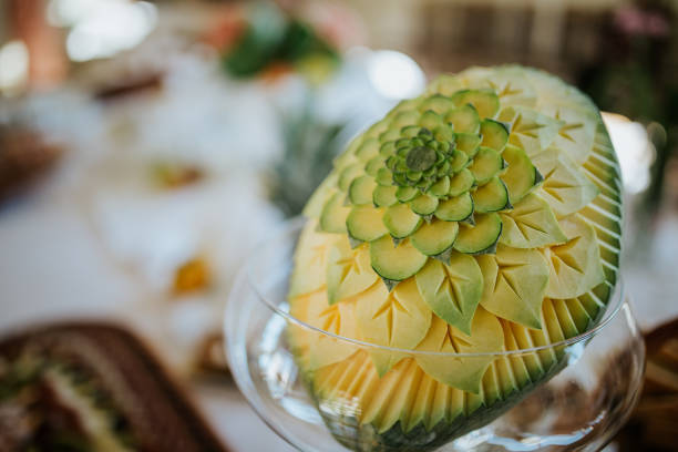 A close-up photo of a beautifully carved watermelon placed in a glass bowl. The watermelon has been intricately carved with a floral pattern, creating a delicate and artistic display. The watermelon is a vibrant red color, and the petals of the flower are green. The background is blurred, suggesting the watermelon is part of a table setting at a special event.