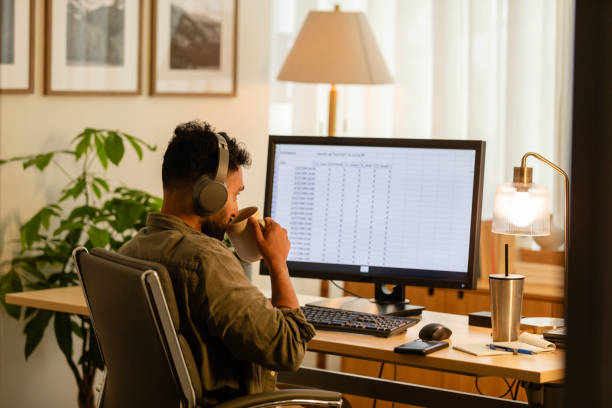 man working in home office sips coffee while reviewing spreadsheet - evden çalışma stok fotoğraflar ve resimler