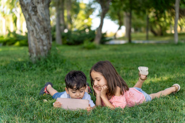 Boy and girl lying on grass using tablet.  Boy and girl watching video on tablet outdoors. Primary school students on summer vacation.