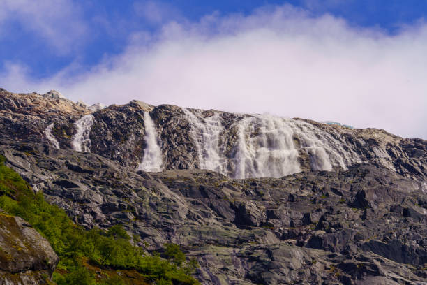 A stunning view of a powerful waterfall descending from a glacier, cascading over rugged rocks under a bright blue sky in the Norwegian mountains