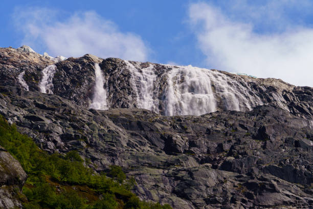 A stunning view of a powerful waterfall descending from a glacier, cascading over rugged rocks under a bright blue sky in the Norwegian mountains