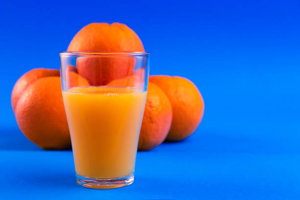 A group of fresh oranges accompanied by a glass of freshly squeezed orange juice on a vibrant blue background. The striking contrast between the oranges, juice, and the vivid blue backdrop creates a visually appealing composition, ideal for use in health and wellness promotions, food and beverage marketing, or bright and fresh product designs